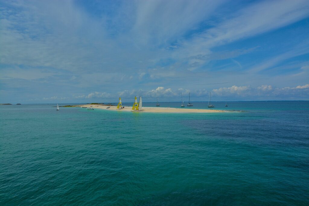 Eine lange weiße Sandzunge, auf der Katamarane mit gelben und weißen Segeln liegen, umgeben von ankernden Segelbooten auf türkisem Wasser, unter blauem Himmel mit Wolkenstreifen.