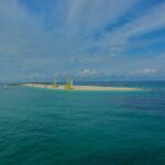 Eine lange weiße Sandzunge, auf der Katamarane mit gelben und weißen Segeln liegen, umgeben von ankernden Segelbooten auf türkisem Wasser, unter blauem Himmel mit Wolkenstreifen.