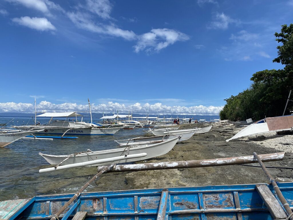 Eine Reihe weißer Bangkas an einer felsigen Küste festgemacht, kleine Auslegerkanus auf den Fels gezogen und ein blaues Boot mit abblätternder Farbe im Vordergrund, unter blauem Himmel mit weißen Wolken.