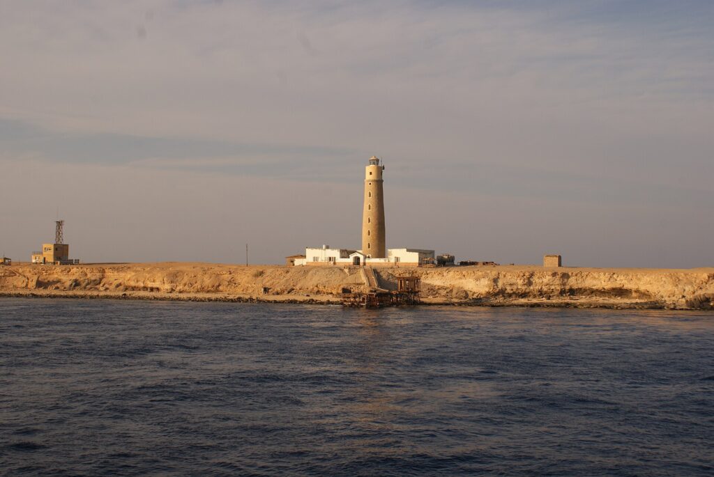 Der Leuchtturm von Big Brother und seine weißen Gebäude auf der Wüsteninsel, im Vordergrund ein rostiger Steg, im goldenen Abendlicht