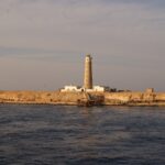 Der Leuchtturm von Big Brother und seine weißen Gebäude auf der Wüsteninsel, im Vordergrund ein rostiger Steg, im goldenen Abendlicht