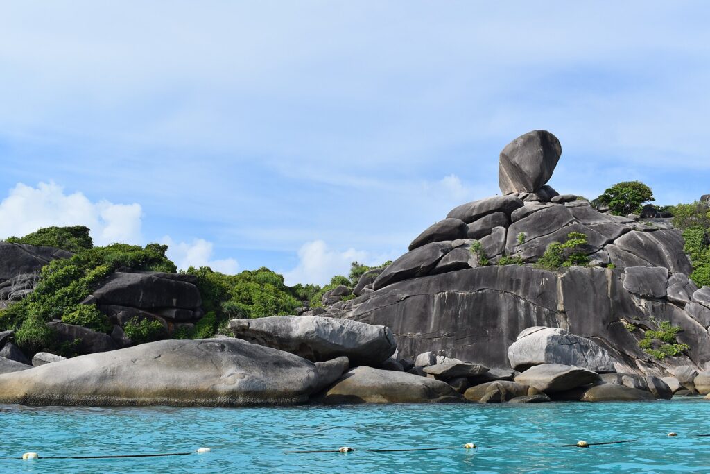 Der riesige, balancierende Granitblock der Insel 8 krönt einen Haufen glatter grauer Felsen am Rand von türkisfarbenem Wasser, unter leicht dunstigem blauem Himmel, im Vordergrund eine Bojenleine
