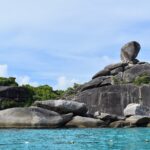 Der riesige, balancierende Granitblock der Insel 8 krönt einen Haufen glatter grauer Felsen am Rand von türkisfarbenem Wasser, unter leicht dunstigem blauem Himmel, im Vordergrund eine Bojenleine