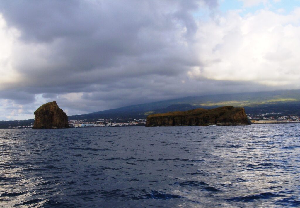 Von einem Boot auf dunkler, bewegter See aus gesehen: links eine aufragende Felsinsel, rechts eine lange flache Insel, dahinter die Stadt Madalena und die Hänge des Pico unter bedecktem Himmel
