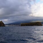 Von einem Boot auf dunkler, bewegter See aus gesehen: links eine aufragende Felsinsel, rechts eine lange flache Insel, dahinter die Stadt Madalena und die Hänge des Pico unter bedecktem Himmel