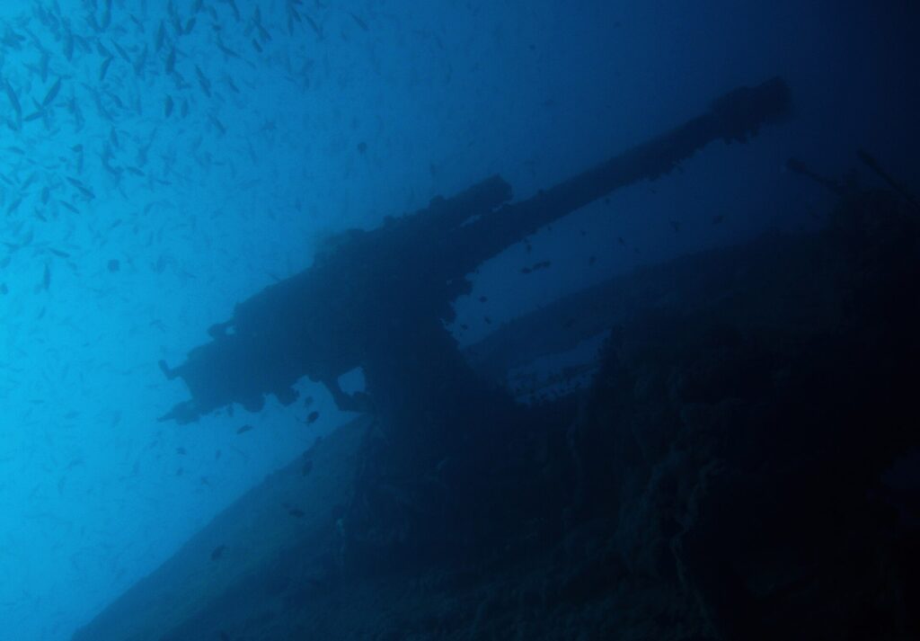 Das Heckgeschütz der Thistlegorm als Silhouette vor dem Blau, unter einem Schwarm kleiner Fische