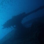 Das Heckgeschütz der Thistlegorm als Silhouette vor dem Blau, unter einem Schwarm kleiner Fische