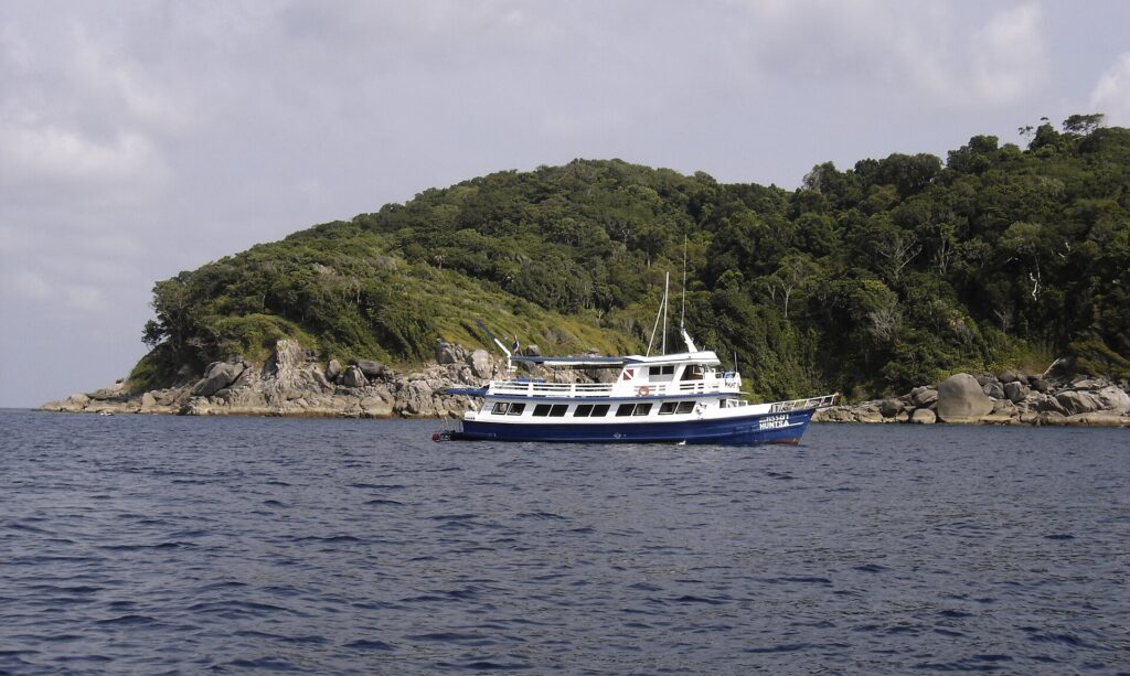 Ein blau-weißes Safariboot ankert vor einer Similan-Insel, deren graue Granitblöcke unter dem Wald bis ans Wasser reichen