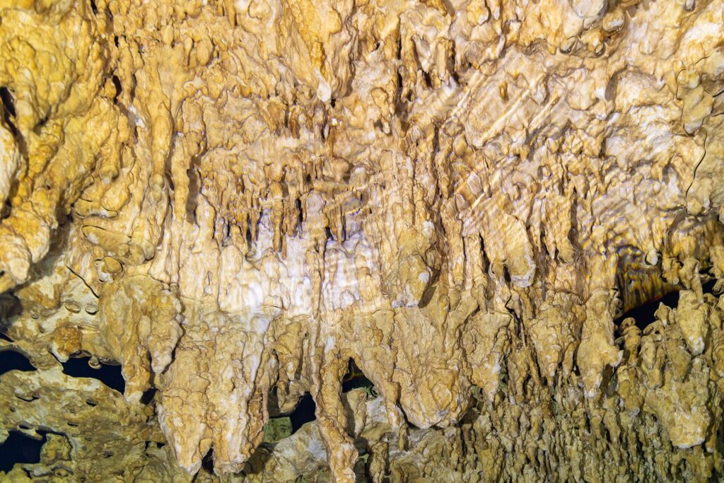 Nahaufnahme hellgelber Tropfsteine und feiner Stalaktiten, unten die gewellte Spiegelung des Wassers
