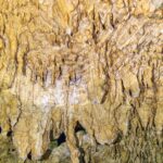 Nahaufnahme hellgelber Tropfsteine und feiner Stalaktiten, unten die gewellte Spiegelung des Wassers