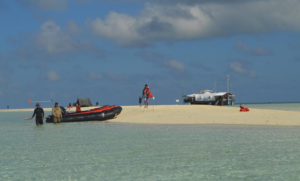 Eine weiße Sandbank in klarem, hellgrünem Wasser, an der gerade ein schwarz-oranges Schlauchboot angelegt hat, Menschen in Schwimmwesten auf dem Sand und rechts die Rangerstation auf Pfählen.