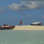 Eine weiße Sandbank in klarem, hellgrünem Wasser, an der gerade ein schwarz-oranges Schlauchboot angelegt hat, Menschen in Schwimmwesten auf dem Sand und rechts die Rangerstation auf Pfählen.