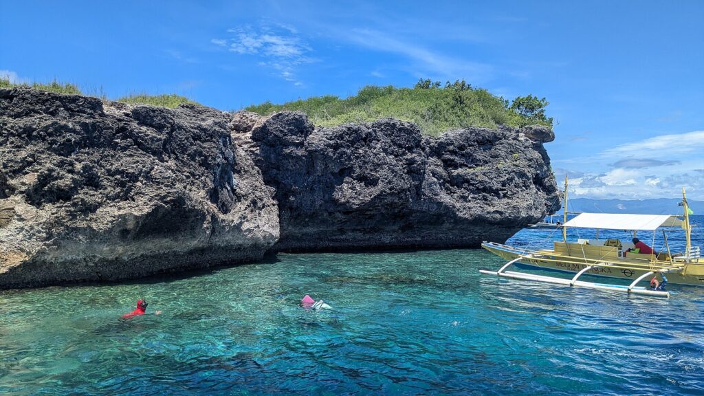 Die graue, ausgehöhlte Kalksteinklippe der Insel Pescador, zwei Schnorchler mit Maske und eine gelbe Banca in türkisfarbenem Wasser