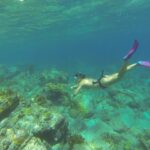 Eine Apnoetaucherin mit Maske und rosa Flossen taucht unter der gekräuselten Oberfläche zu einem Grund aus Felsen und Korallen ab, in türkisem Wasser