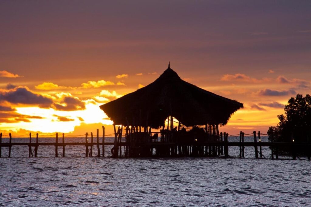 Der Pavillon der Fundu Lagoon mit Strohdach, auf Pfählen am Ende des Stegs, bei Sonnenuntergang