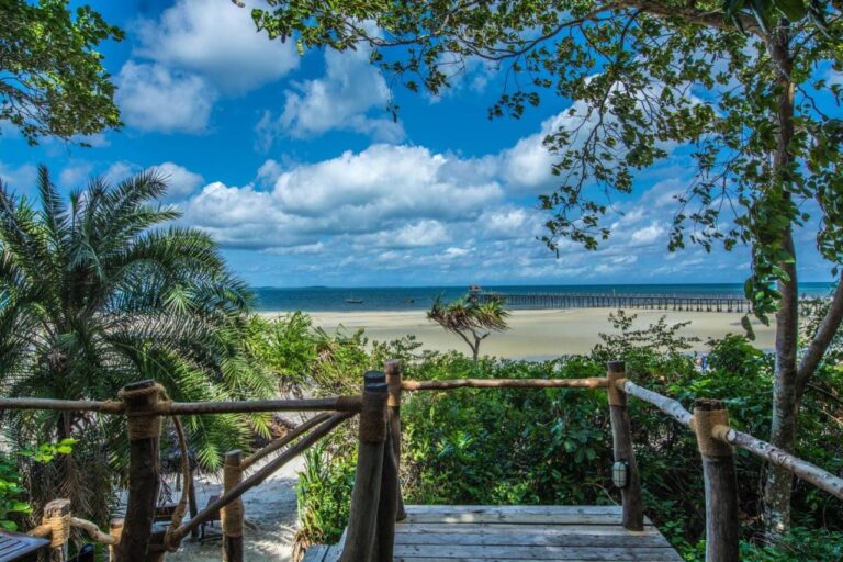 Blick von einer Holzterrasse auf einen breiten Sandstrand und einen langen Steg