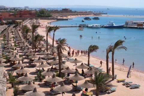 Der Strand des Giftun Azur Resorts mit Strohschirmen und Palmen, im Hintergrund ein Steg und Boote