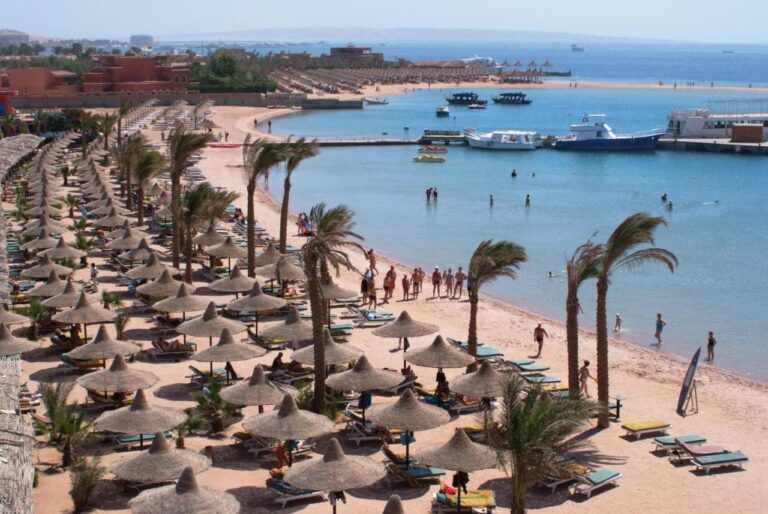 Der Strand des Giftun Azur Resorts mit Strohschirmen und Palmen, im Hintergrund ein Steg und Boote