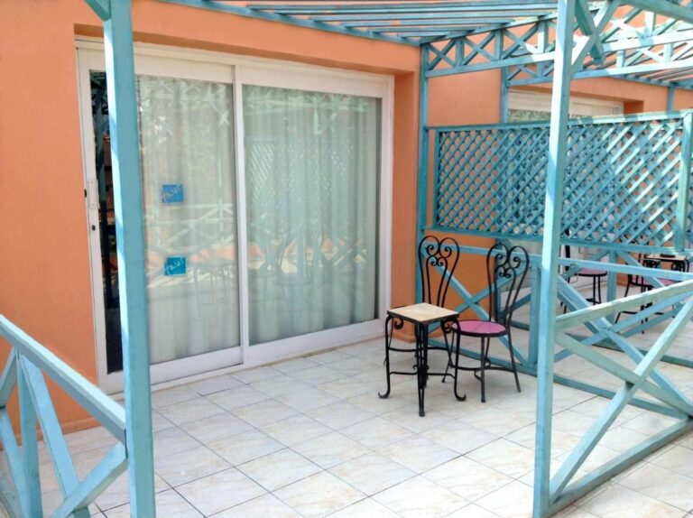 Eine geflieste Terrasse unter einer türkisfarbenen Pergola, mit kleinem Tisch und zwei Stühlen aus Schmiedeeisen