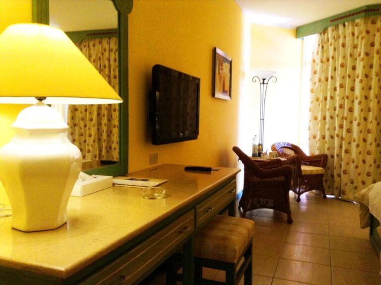 Ein Zimmer mit gelben Wänden, Schreibtisch, Fernseher und zwei Rattansesseln am Fenster