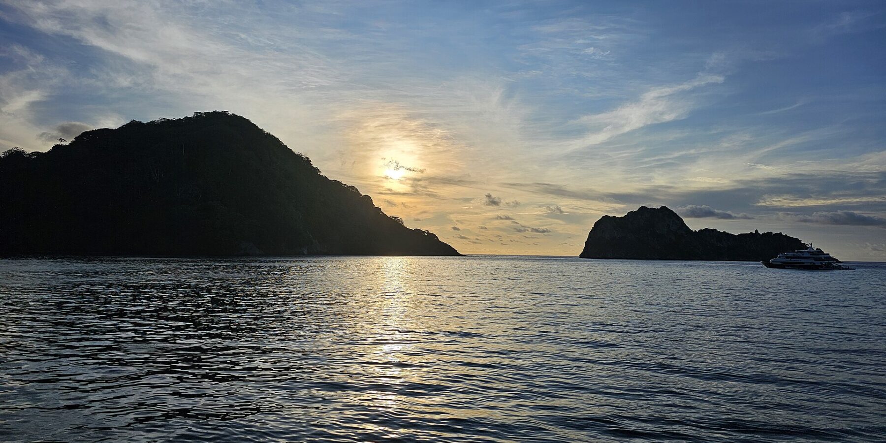 Die Kokosinsel im Gegenlicht bei Sonnenuntergang, rechts ein ankerndes Tauchsafarischiff, ruhige See