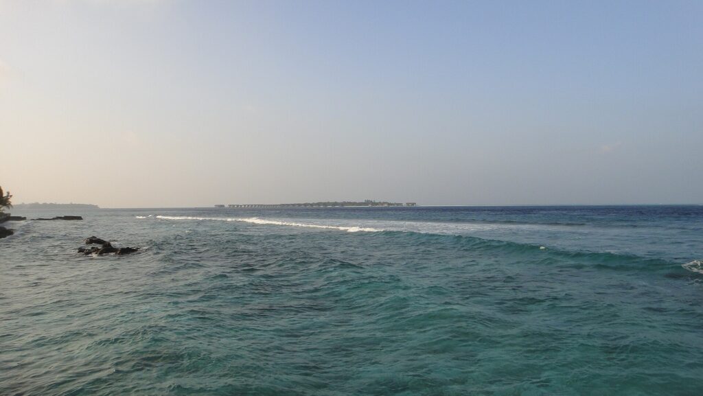 Von einem felsigen Ufer aus ein bewegter türkiser Kanal, in dem eine Wellenlinie über das Riff bricht, am Horizont eine flache Insel mit Wasserbungalows unter blassem Himmel.