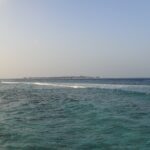 Von einem felsigen Ufer aus ein bewegter türkiser Kanal, in dem eine Wellenlinie über das Riff bricht, am Horizont eine flache Insel mit Wasserbungalows unter blassem Himmel.