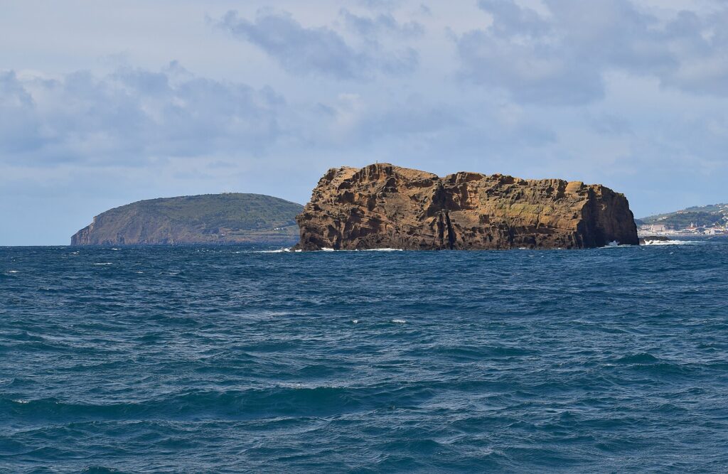 Das Ilhéu Deitado, ein langer Block aus ockerfarbenem Tuff mit wabenartig ausgehöhlten Klippen, auf bewegtem dunkelblauem Meer, dahinter der grüne Hügel des Monte da Guia und rechts in der Ferne Häuser auf Faial