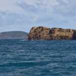 Das Ilhéu Deitado, ein langer Block aus ockerfarbenem Tuff mit wabenartig ausgehöhlten Klippen, auf bewegtem dunkelblauem Meer, dahinter der grüne Hügel des Monte da Guia und rechts in der Ferne Häuser auf Faial
