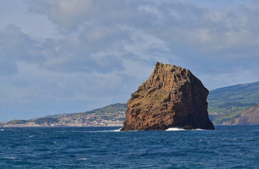 Das Ilhéu em Pé, ein hoher ockerfarbener Tufffelsen mit ausgewaschenen Schichten, ragt aus dunkelblauem Meer mit ein paar brechenden Wellen, vor der weißen Stadt Horta und den grünen Hügeln von Faial unter bewölktem Himmel