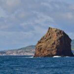 Das Ilhéu em Pé, ein hoher ockerfarbener Tufffelsen mit ausgewaschenen Schichten, ragt aus dunkelblauem Meer mit ein paar brechenden Wellen, vor der weißen Stadt Horta und den grünen Hügeln von Faial unter bewölktem Himmel