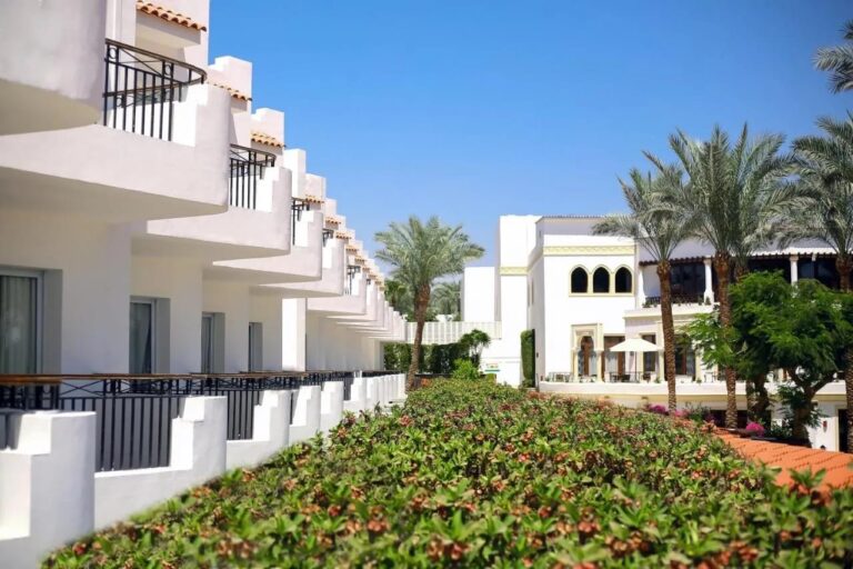 Die weißen Gebäude des JAZ Fanara Resorts mit ihren Balkonen und Palmen, hinter einer blühenden Hecke