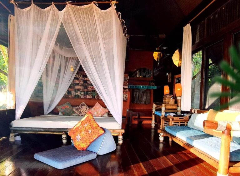 Ein Cottage mit einem Bett unter einem Moskitonetz, einer Bambusbank mit blauen Kissen und dunklem Parkett