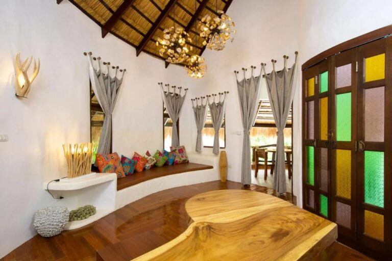 Ein weißer Wohnraum mit einer Sitzbank mit bunten Kissen, einem Massivholztisch und Türen mit Buntglas