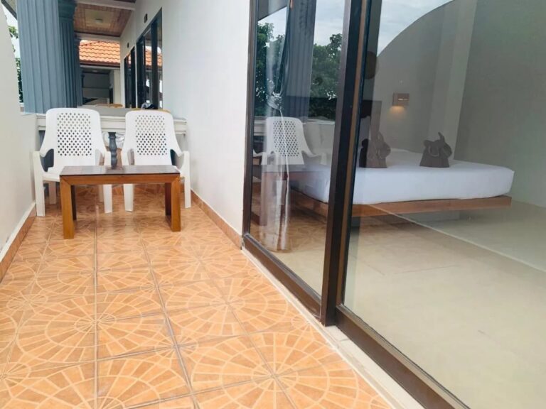 Ein gefliester Balkon mit zwei weißen Stühlen und einem Couchtisch, vor der Fensterfront eines Zimmers