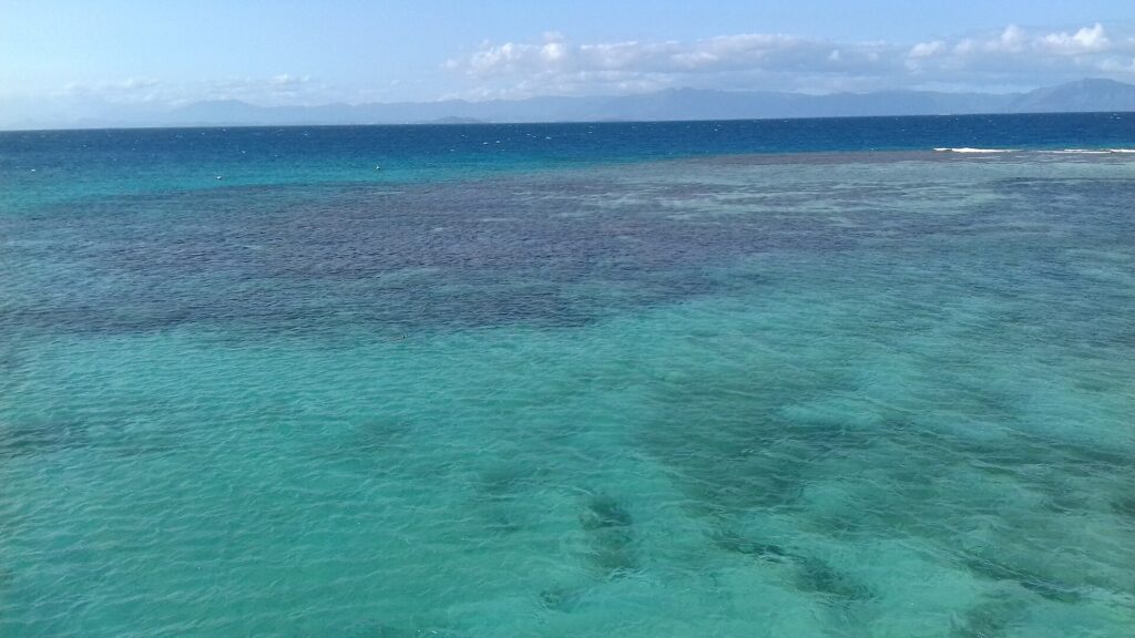 Die Lagune von oben, türkisfarbenes Wasser mit dunklen Korallenblöcken, rechts das Barriereriff mit brechenden Wellen und am Horizont die Berge der Grande Terre
