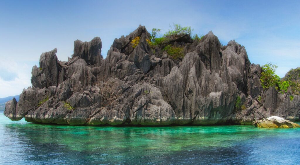 Eine graue Kalksteininsel voller Spitzen, mit etwas Vegetation obenauf, ragt über klarem, türkisfarbenem Wasser auf, in dem die Korallen zu erahnen sind, unter blauem Himmel.