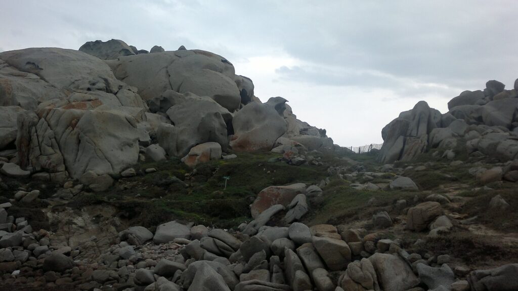 Große, runde, von der Erosion ausgehöhlte graue Granitblöcke mit orangefarbenen Flechten rund um eine kurze Wiese, auf der ein Pfad mit Holzzaun zum Meer führt, unter bedecktem Himmel.