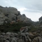 Große, runde, von der Erosion ausgehöhlte graue Granitblöcke mit orangefarbenen Flechten rund um eine kurze Wiese, auf der ein Pfad mit Holzzaun zum Meer führt, unter bedecktem Himmel.