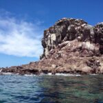 Die rötlichen Felsen von Los Islotes unter blauem Himmel, mit Seelöwen, die knapp über dem Wasser liegen, und einer zerklüfteten Klippe darüber