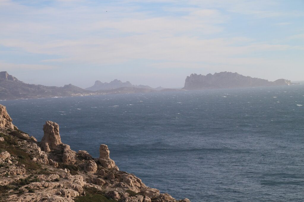 Von den Kalkfelsen des Frioul im Vordergrund geht der Blick über ein raues Meer mit Schaumkronen bis zur Île Maïre und zur Küste von Les Goudes, die sich im Dunst abzeichnen.