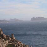 Von den Kalkfelsen des Frioul im Vordergrund geht der Blick über ein raues Meer mit Schaumkronen bis zur Île Maïre und zur Küste von Les Goudes, die sich im Dunst abzeichnen.