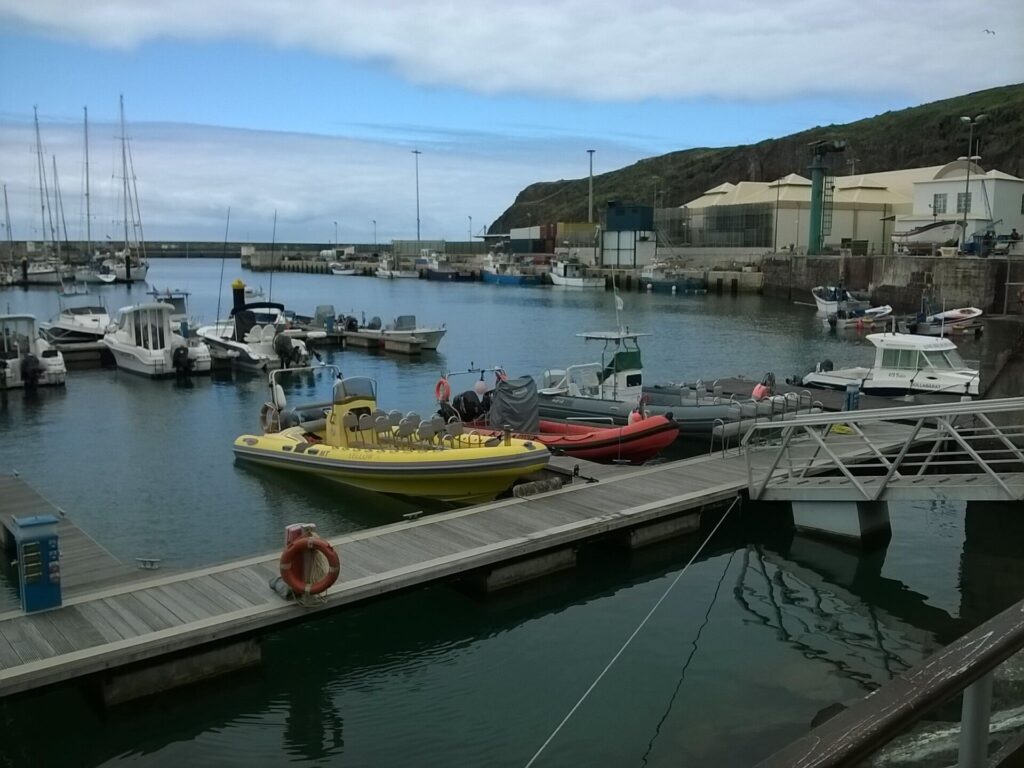 Schlauchboote, ein gelbes mit Sitzreihen und ein rotes, am Steg der Marina von Vila do Porto festgemacht, dahinter Segel- und Fischerboote unter einer grünen Klippe und bewölktem Himmel