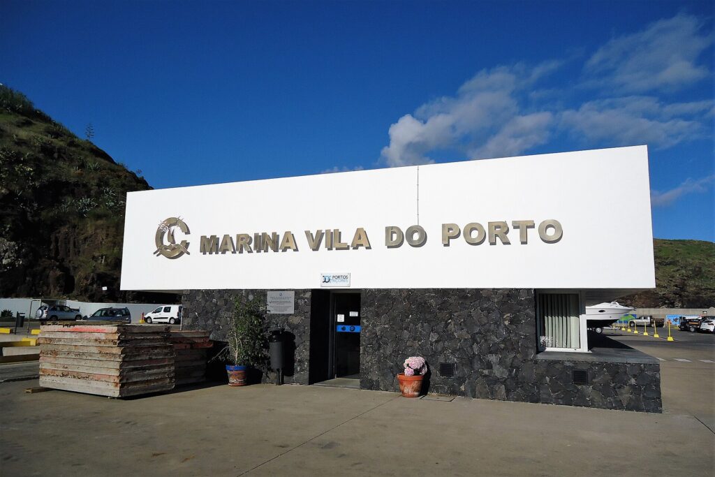 Das Empfangsgebäude der Marina von Vila do Porto, großes weißes Schild mit Metallbuchstaben an einer schwarzen Basaltmauer, unter blauem Himmel, links eine grüne Klippe