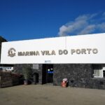 Das Empfangsgebäude der Marina von Vila do Porto, großes weißes Schild mit Metallbuchstaben an einer schwarzen Basaltmauer, unter blauem Himmel, links eine grüne Klippe