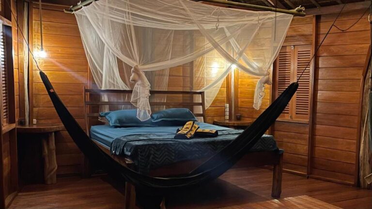 Ein Zimmer mit Holzwänden im Lampenlicht, mit einem Bett unter einem Moskitonetz und einer schwarzen Hängematte