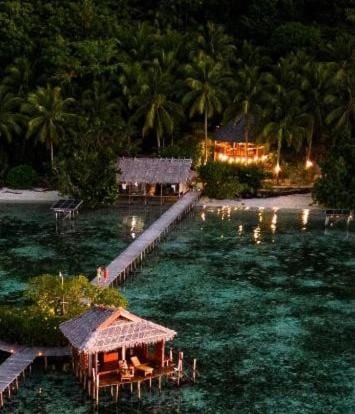 Das Nyande Raja Ampat in der Abenddämmerung aus der Luft, mit einem Bungalow auf Pfählen und dem Steg zum Strand