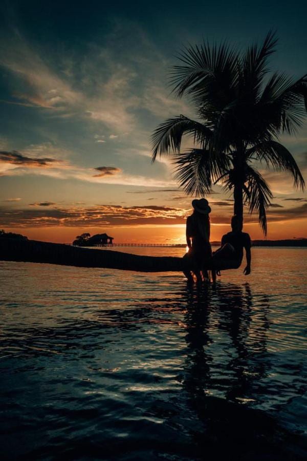 Zwei Silhouetten auf dem schrägen Stamm einer Palme über dem Meer, unter einem orangefarbenen Himmel