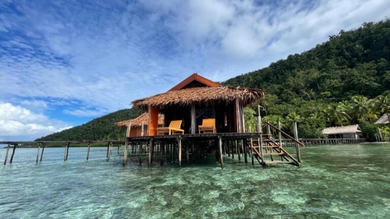 Ein Bungalow des Nyande Raja Ampat auf Pfählen über klarem Wasser, am Fuß eines bewaldeten Hügels