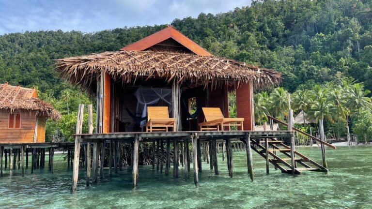 Ein Bungalow auf Pfählen mit Palmdach, zwei Liegen und einer Treppe, die ins Wasser führt